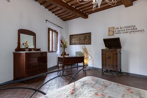 Quarto com cômoda de madeira, escrivaninha com livros, espelho, cama com colcha florida e parede com uma frase.