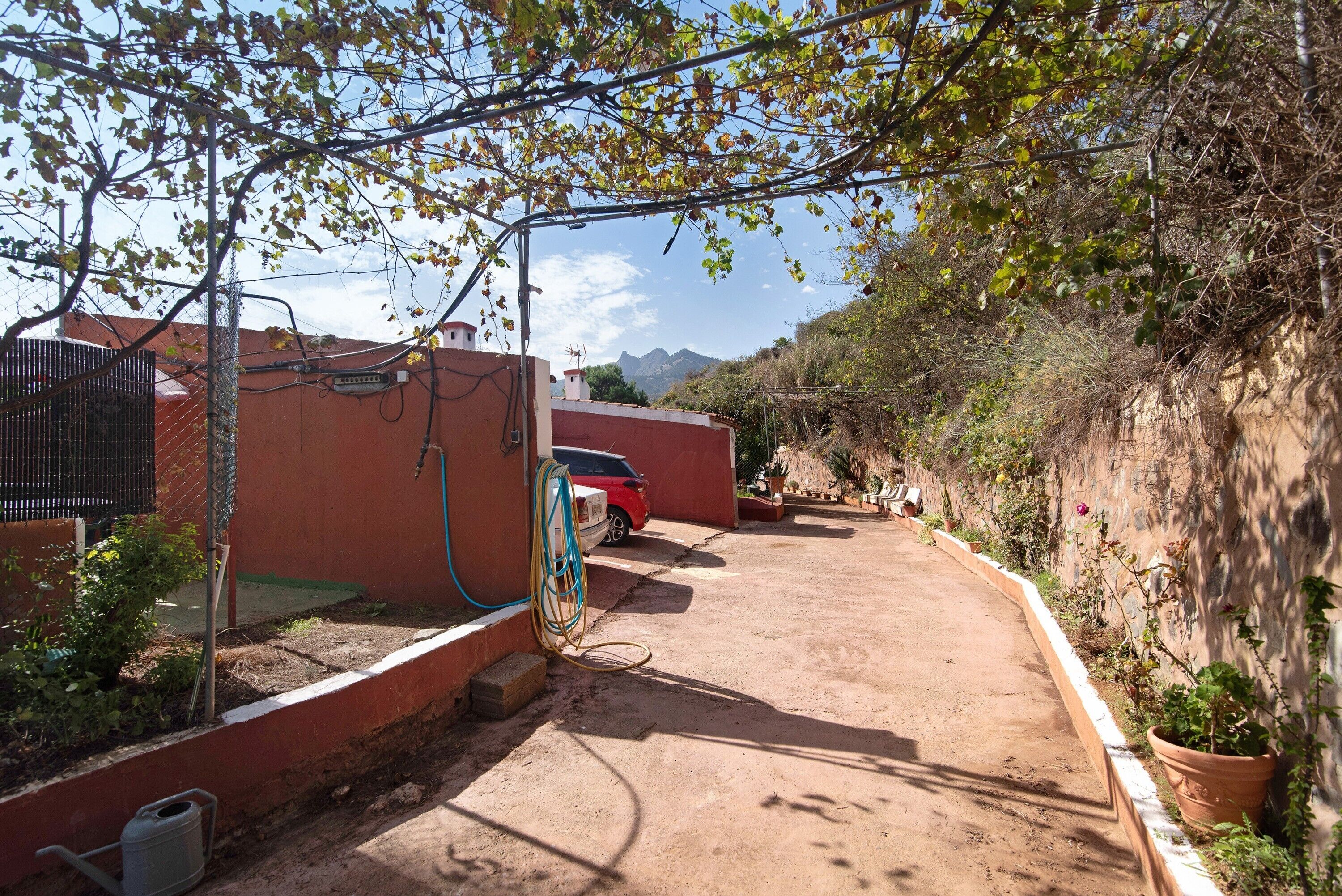 Un chemin de terre mène à un bâtiment rouge, devant lequel est garée une voiture rouge. L'ensemble est entouré de végétation et se trouve sur fond de montagnes.
