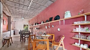 A room with wooden chairs and tables, a wall-mounted shelf with hats, and a table with a small potted plant.