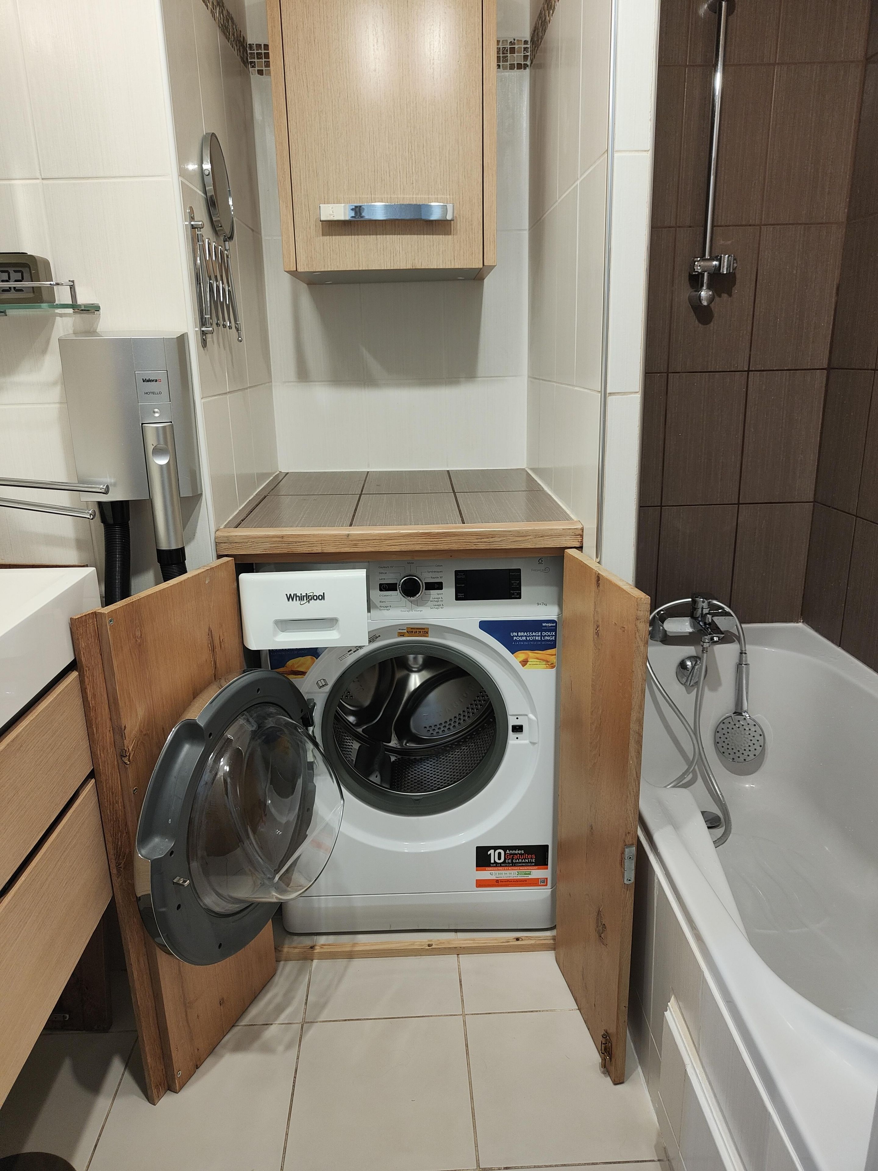 A compact hotel bathroom with a built-in washer and dryer, a shower, and a wooden cabinet.
