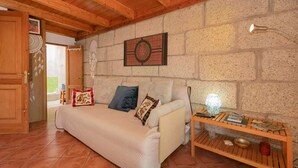 Um quarto acolhedor com teto de madeira, parede de pedra, sofá com almofadas, pequena mesa com livros e uma luminária, e uma porta que dá para o exterior.