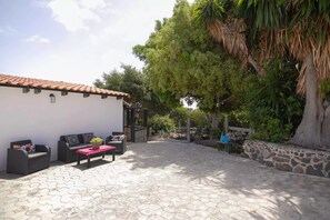 A patio area with a tiled floor, outdoor furniture, a white building with a tiled roof, and a large tree.