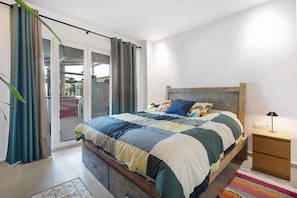 Un dormitorio con cama, mesita de noche, ventana con cortinas y vistas al exterior.