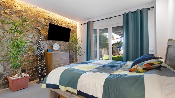 Un dormitorio con pared de piedra, televisor, cama con colcha y vistas al exterior.
