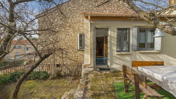 A stone building with a wooden bench and table outside, a door, and a window with shutters.
