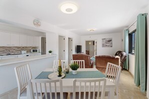 Een moderne keuken met witte kasten, een eethoek met witte stoelen, een tafel met een groene placemat en een bruine bank in de woonkamer.
