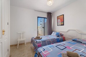 Een slaapkamer met twee bedden, een witte stoel, een deur en een schilderij aan de muur.