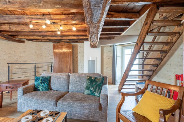 Un interior rústico con vigas de madera a la vista, un sofá con cojines estampados, una silla de madera y una escalera.