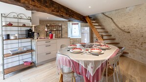 A dining area with a table set for four, a kitchen with a wooden beam ceiling, and a staircase. - La Tourette – Charming, Unique Cottage with Private Terrace, Garden, and Wi-Fi (Jard Sur Mer)