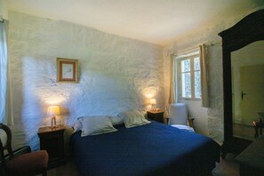 Un dormitorio con una cama, dos mesitas de noche con lámparas, una silla, una ventana con cortinas y un cuadro en la pared.