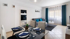 Un comedor con una mesa de vidrio dispuesta para cuatro personas, un sofá, un televisor y una ventana con cortinas.