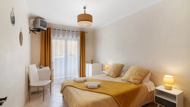 Un dormitorio con cama, mesita de noche, lámpara, silla y televisor montado en la pared.