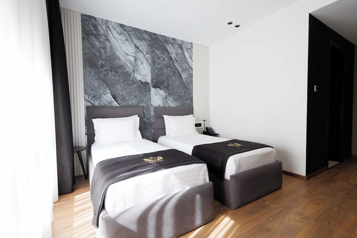 A modern hotel room with two beds, marble wall, wooden flooring, and a black door.