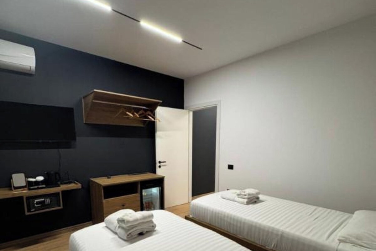 A modern hotel room with two single beds, a desk with a chair, a small table, and a built-in TV.