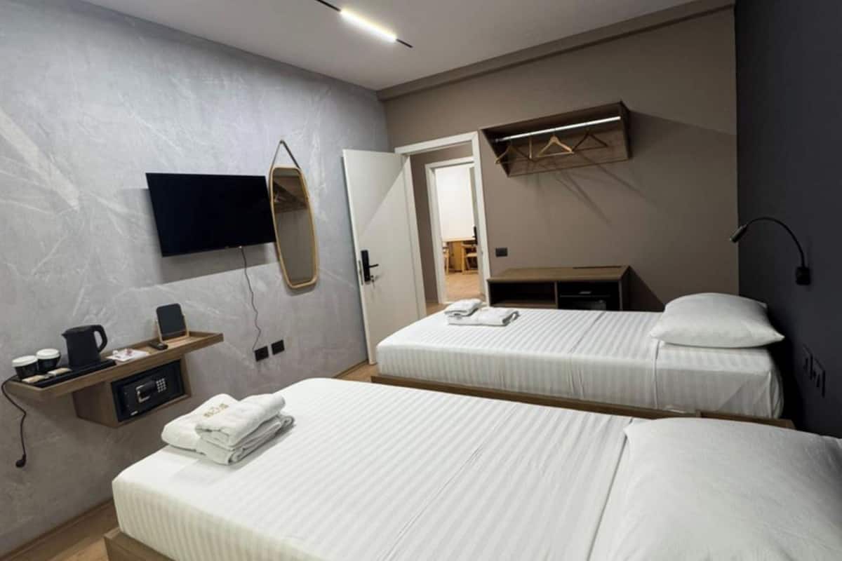 A modern hotel room with two beds, a flat-screen TV mounted on the wall, a wooden bedside table with a kettle and cups, and a small shelf with a phone and a coffee maker.