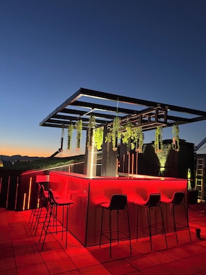 An outdoor bar area with a red-lit bar counter, hanging plants, and a view of the landscape. - Destiny Hotel (Kashar)