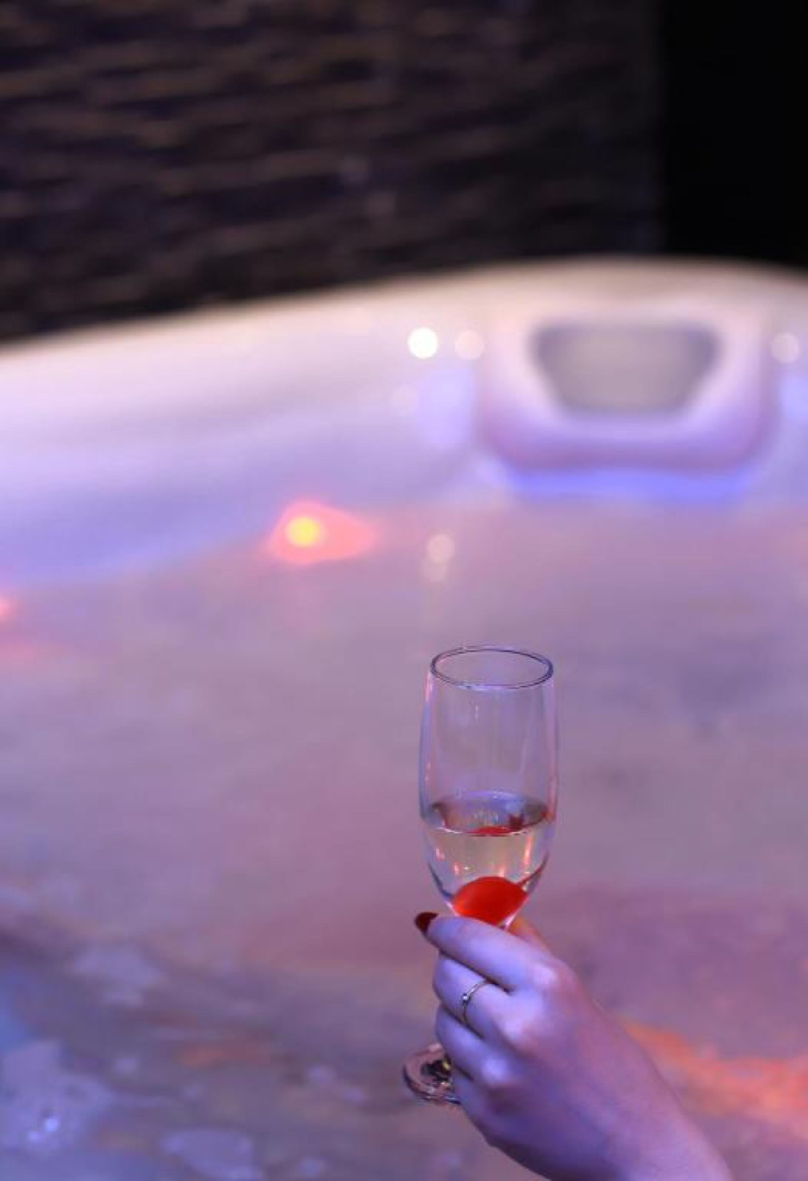 Eine Hand hält ein Glas mit einem roten Objekt, vermutlich ein Cocktail, vor einer beleuchteten Badewanne.