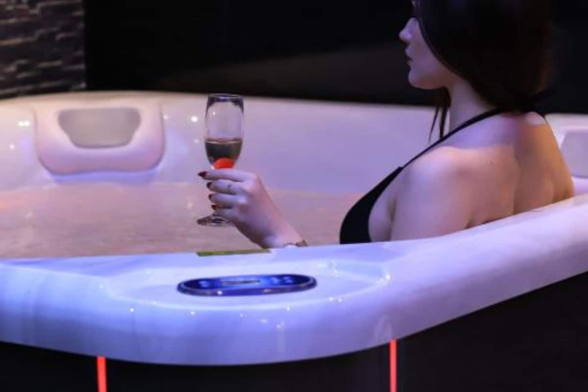 A person relaxing in a hot tub with a glass of wine, surrounded by a dark, tiled wall.