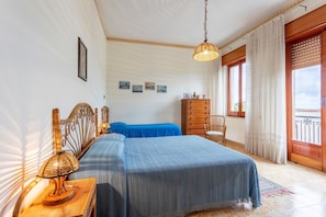 A bedroom with a bed, a bedside table with a lamp, a dresser, a chair, and a window with curtains.