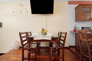 A dining area with a wooden table, chairs, a clock, and a television mounted on the wall.