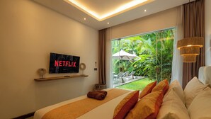 A modern hotel room with a bed, a TV displaying 'NETFLIX', a window with a view of greenery, and a ceiling light fixture. - Flores Villa by Betterplace (Canggu)