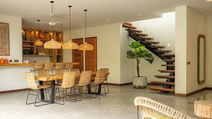 A modern dining area with a wooden table and wicker chairs, a staircase with wooden steps, and a large potted plant. - Flores Villa by Betterplace (Canggu)