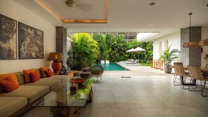 A modern living room with a sofa, coffee table, and chairs, a pool area with lounge chairs, and a view of lush greenery. - Flores Villa by Betterplace (Canggu)