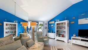 Una sala de estar con sofá, sillones, una mesa de centro y televisor. Se observan estanterías y una planta.