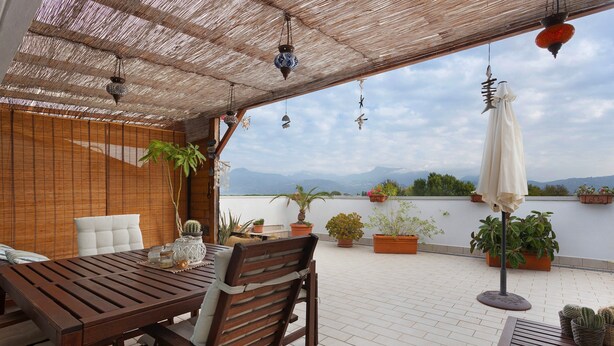 Una mesa y sillas de madera bajo un techo de paja, con plantas en macetas y vistas a las montañas.