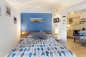 Ein Schlafzimmer mit einem Bett, einem Nachttisch, einer Lampe, einem Gemälde und Blick in die Küche.