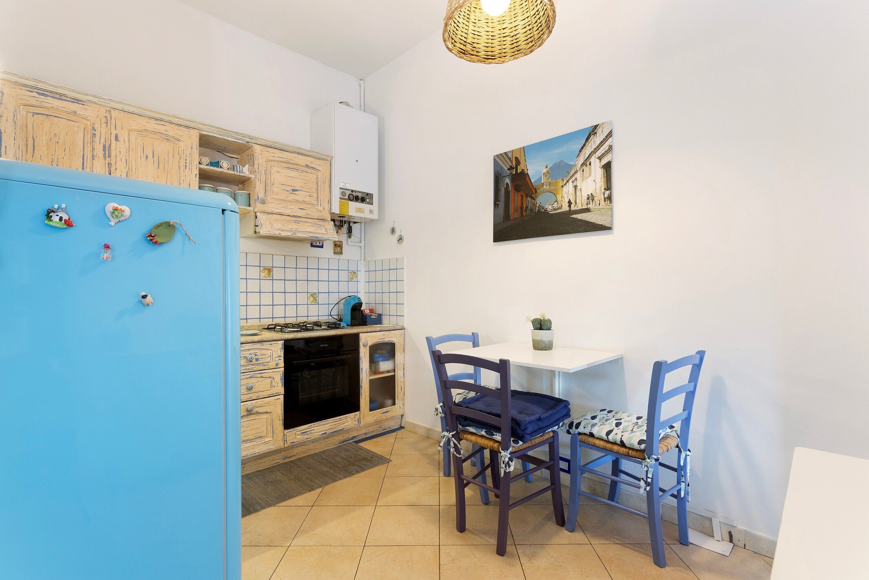 Een keuken met een blauwe koelkast, houten kasten, een betegelde spatwand, een eettafel met stoelen en een aan de muur hangend schilderij.