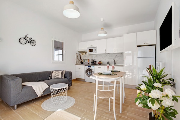 A modern kitchen with white cabinets, a microwave, and a refrigerator. There's a gray sofa with a patterned throw pillow, a small round table with a white chair, and a decorative plant.
