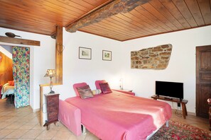 A bedroom with a wooden ceiling, a bed with pink bedding, a television, and a stone wall.