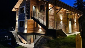 A log cabin with a staircase leading to a deck, surrounded by a stone wall and a mountain backdrop.