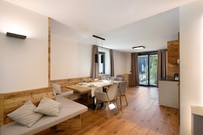 Un interior moderno con una mesa de comedor de madera, un sofá con cojines, una zona de cocina y un ventanal corredero que da a un balcón.