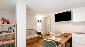 Et moderne, lyst værelse med et træspisebord, en sofa, en bogreol og et vægmonteret fjernsyn.