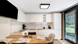 Et moderne køkken med hvide skabe, et spisebord med stole og udsigt til en skov gennem en skydedør.
