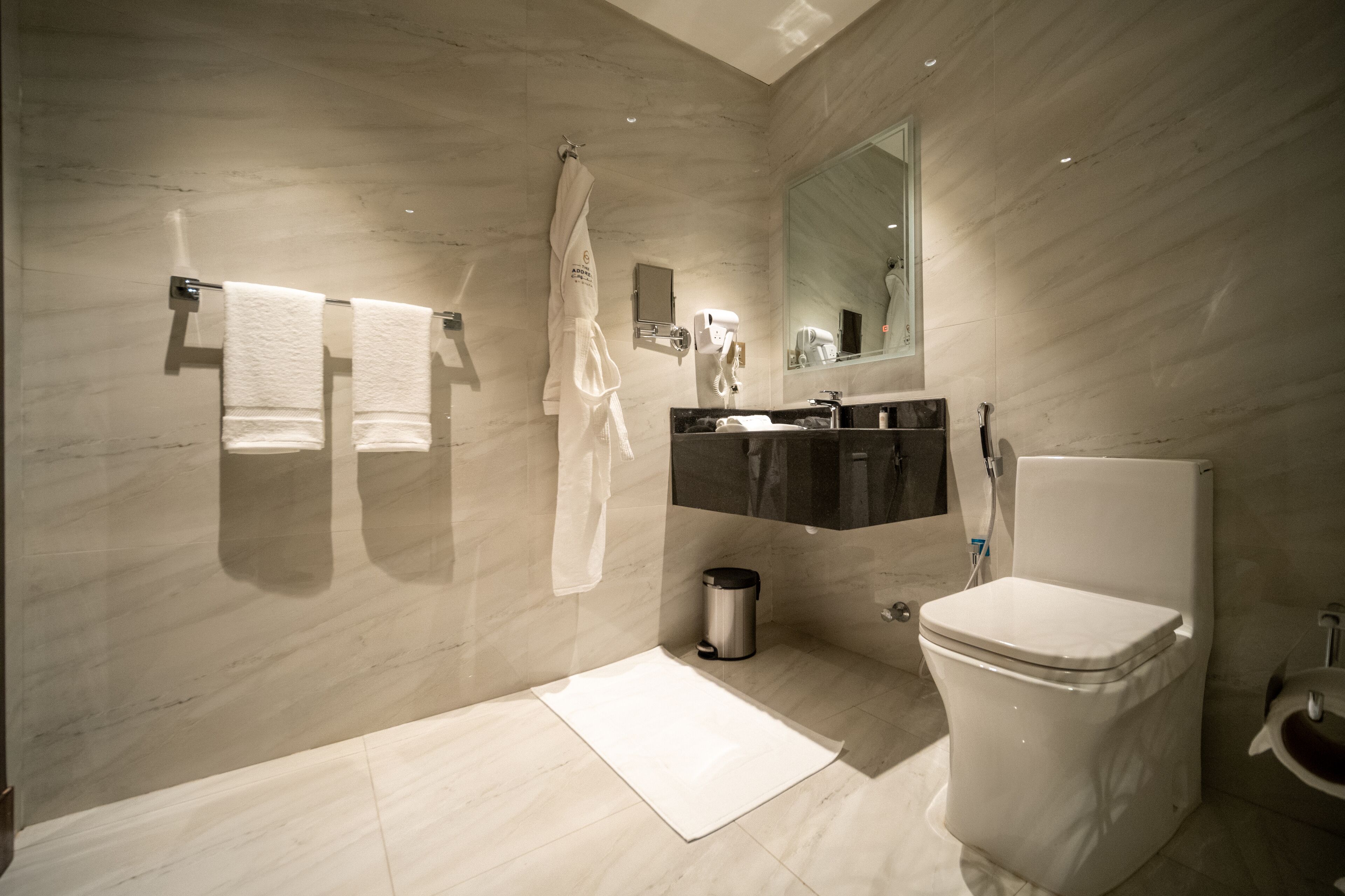 Un baño moderno con piso de mármol, inodoro blanco, tocador negro, espejo y dos toallas colgadas en la pared.