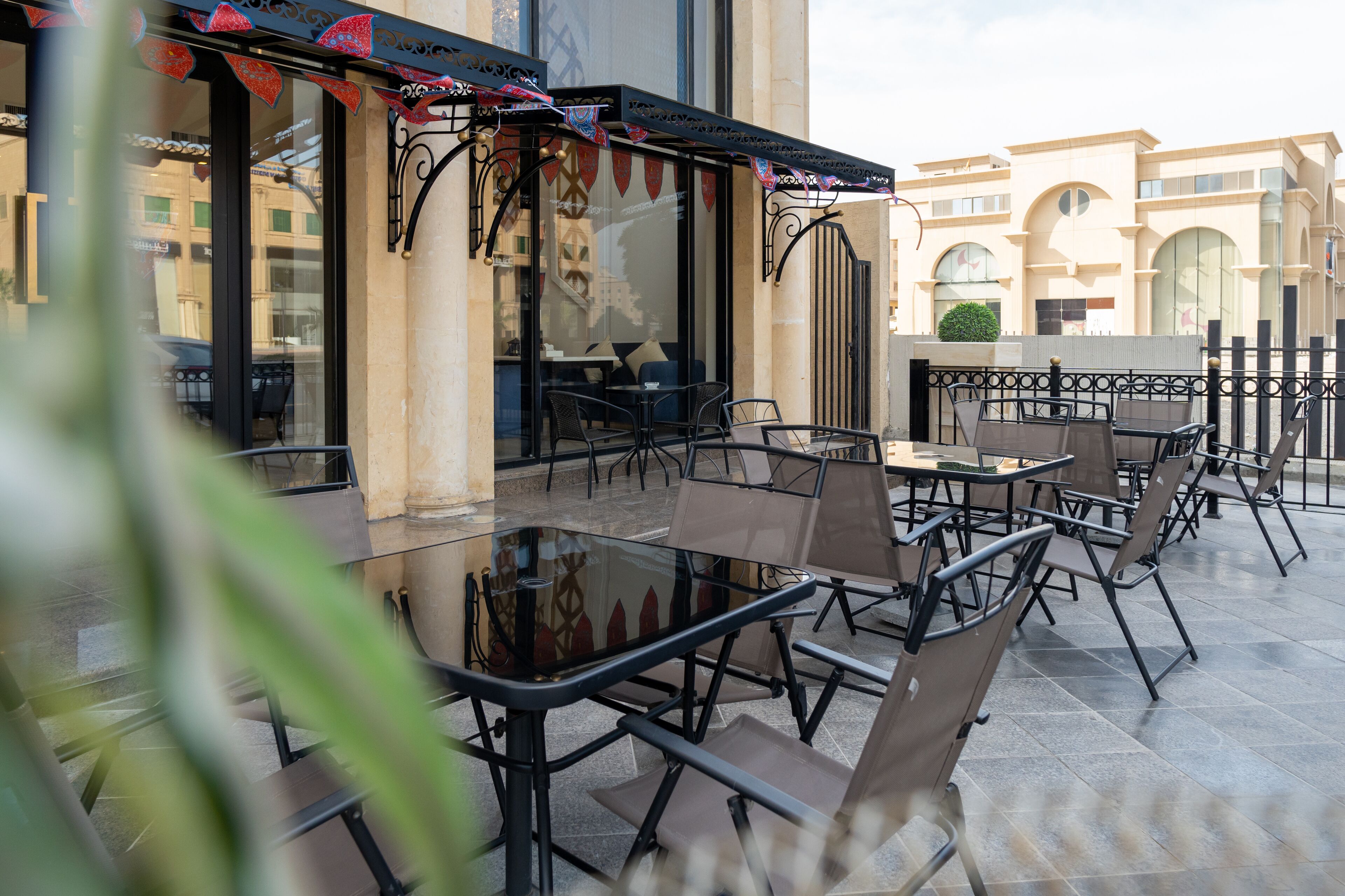 Un café al aire libre con mesas y sillas, una mesa con superficie de vidrio y un toldo decorativo.