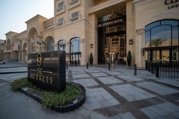 Entrada de um hotel com uma placa indicando "The Address Hotel" e uma porta de entrada grande.