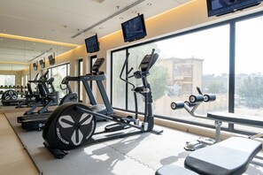 Uma academia com esteiras, elípticos e aparelhos de musculação. Janelas amplas oferecem vista para prédios.