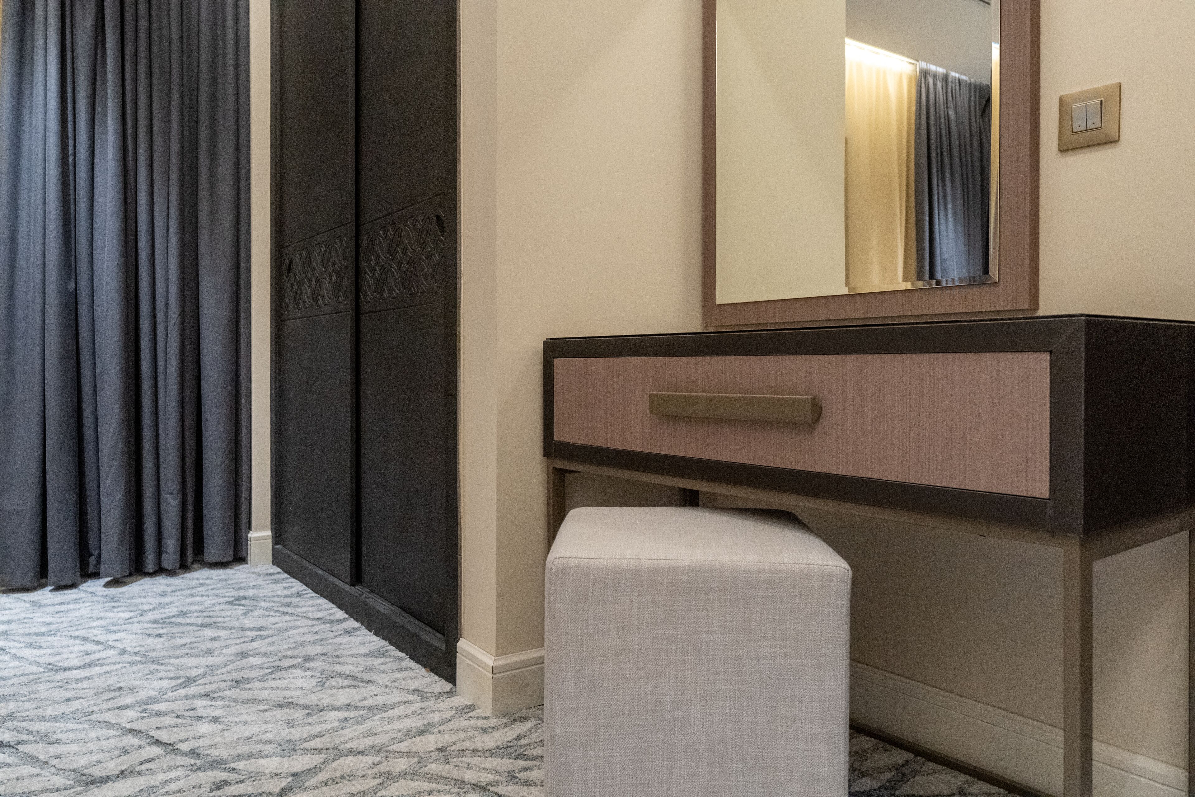 A hotel room with a dark-colored wardrobe, a light-colored bench, a mirror above a wooden vanity, and a patterned carpet.