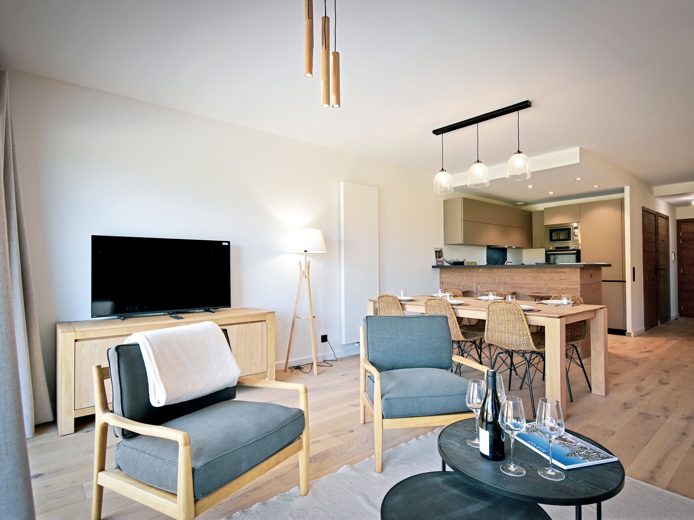 Ein modernes Wohnzimmer mit Flachbildfernseher, zwei Sesseln, einem Esstisch mit Stühlen, einer Küchenzeile und einem kleinen Couchtisch mit Weingläsern und einer Zeitschrift.