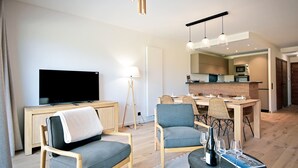 A modern living room with a flat-screen TV, two armchairs, a dining table with chairs, a kitchen area, and a small coffee table with wine glasses and a magazine.