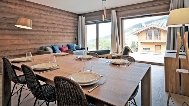 A dining area with a wooden table set for four, surrounded by wicker chairs, with a view of a mountain landscape through large windows.