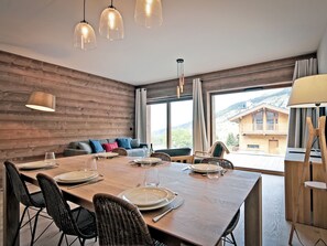 A dining area with a wooden table set for four, surrounded by wicker chairs, with a view of a mountain landscape through large windows. - Spacious apartment with terrace 2 min from slopes, covered parking and concierge service (Méribel)
