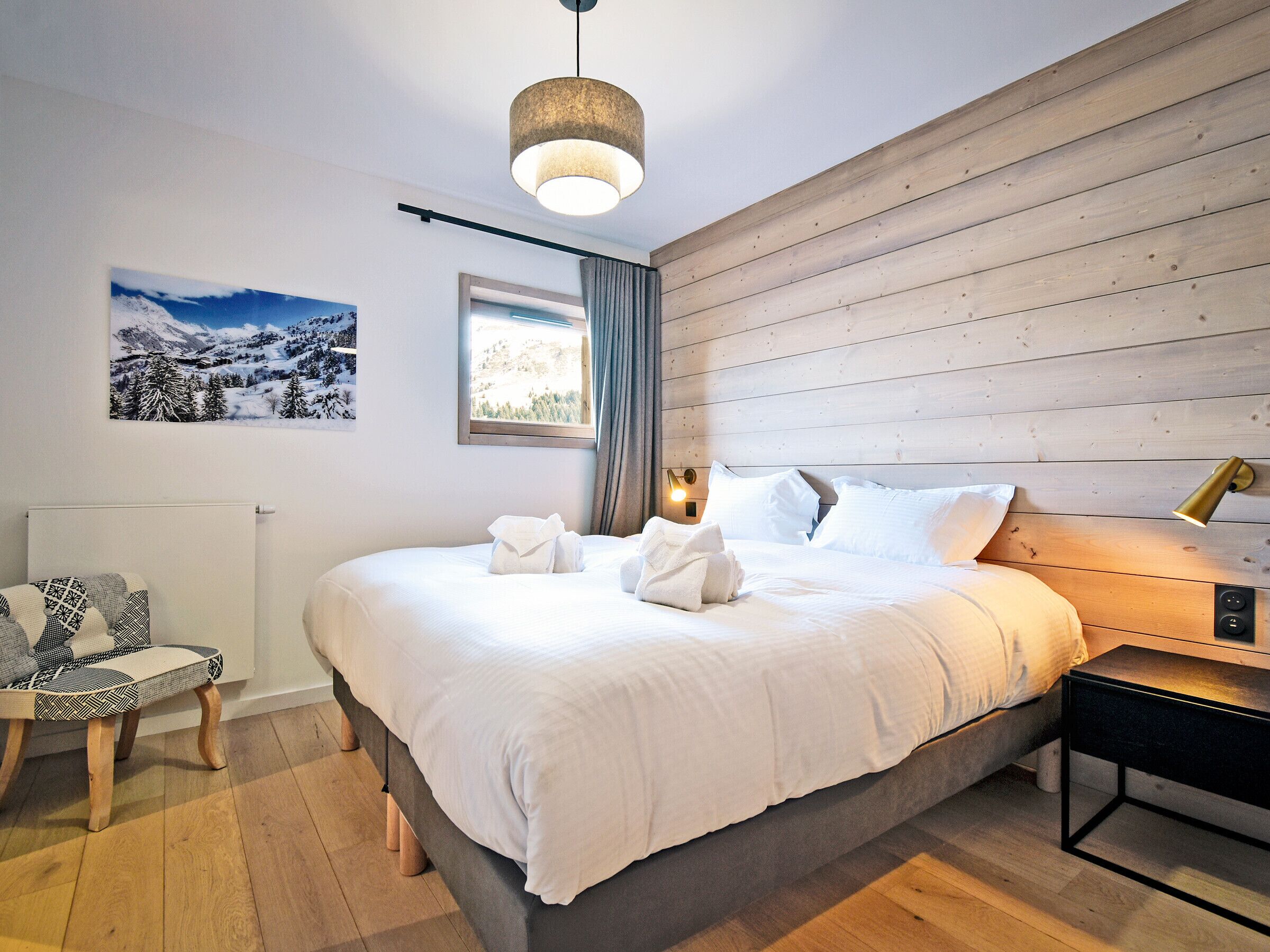 A modern bedroom with a large bed, a bedside table, a chair, a window, and a wall-mounted light fixture.