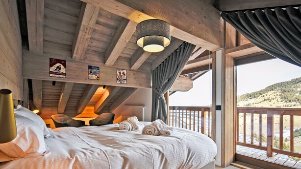 A bedroom with a large bed, wooden beams, a hanging light fixture, and a view of a mountainous landscape through a balcony.