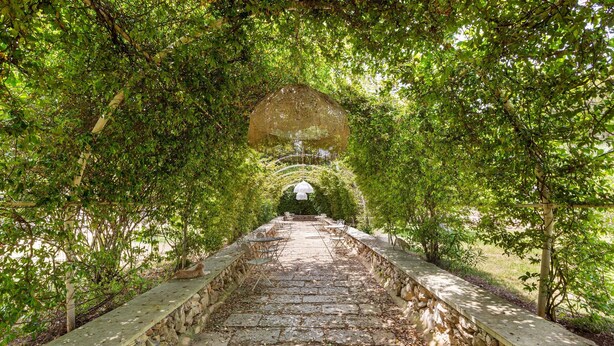 Un sentier de pierres bordé de murs de pierre et d’une végétation luxuriante, qui mène à un belvédère blanc.