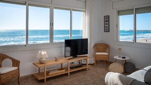 Ett rum med en TV på ett träställ, flätade stolar, ett litet bord och stora fönster med utsikt över havet.